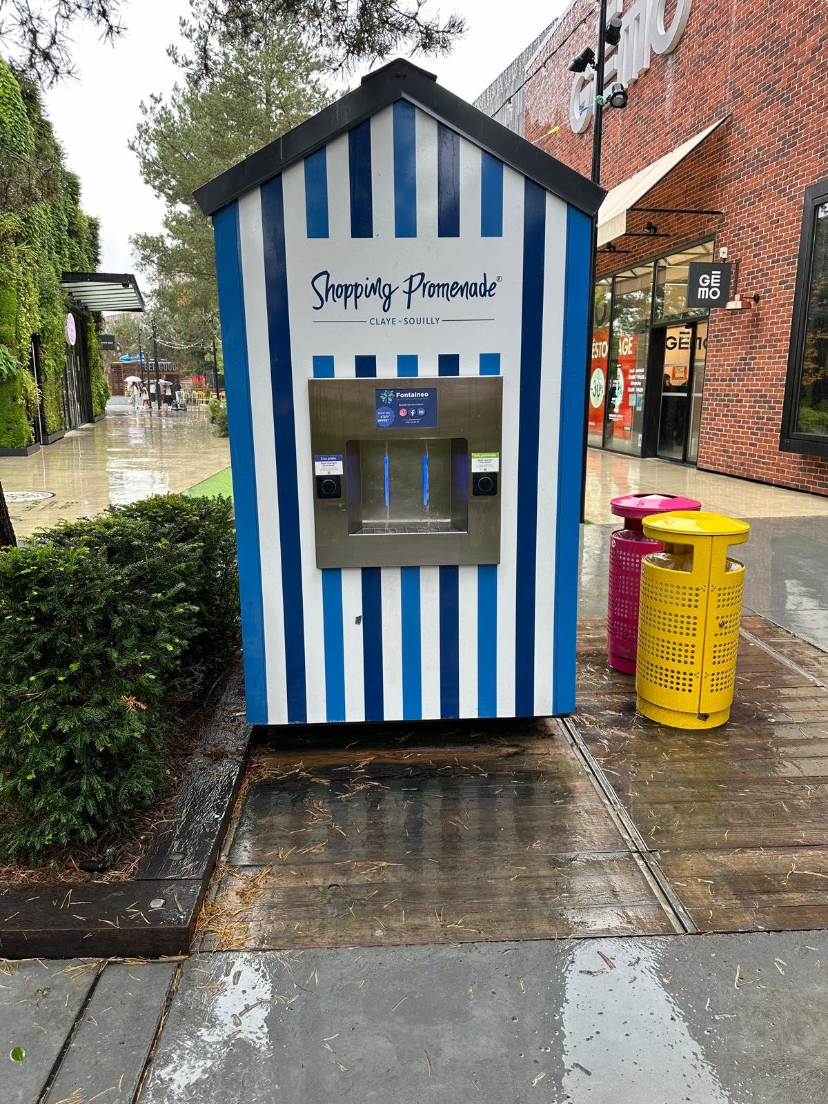 Un distributeur automatique dans un kiosque rayé bleu et blanc sur lequel est écrit « Shopping Promenade » est installé à l'extérieur, sur un trottoir mouillé. Des poubelles aux couleurs vives se trouvent à proximité, et les environs sont humides en raison des pluies récentes.