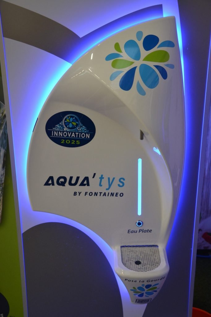 Fontaine à eau blanche avec un logo de Fontaineo et les mots « Aqua'tys by Fontaineo » et « Innovation 2025 ». La fontaine est dotée d'une fente étiquetée « Eau Plate » avec une grille circulaire en dessous, entourée d'un éclairage bleu.