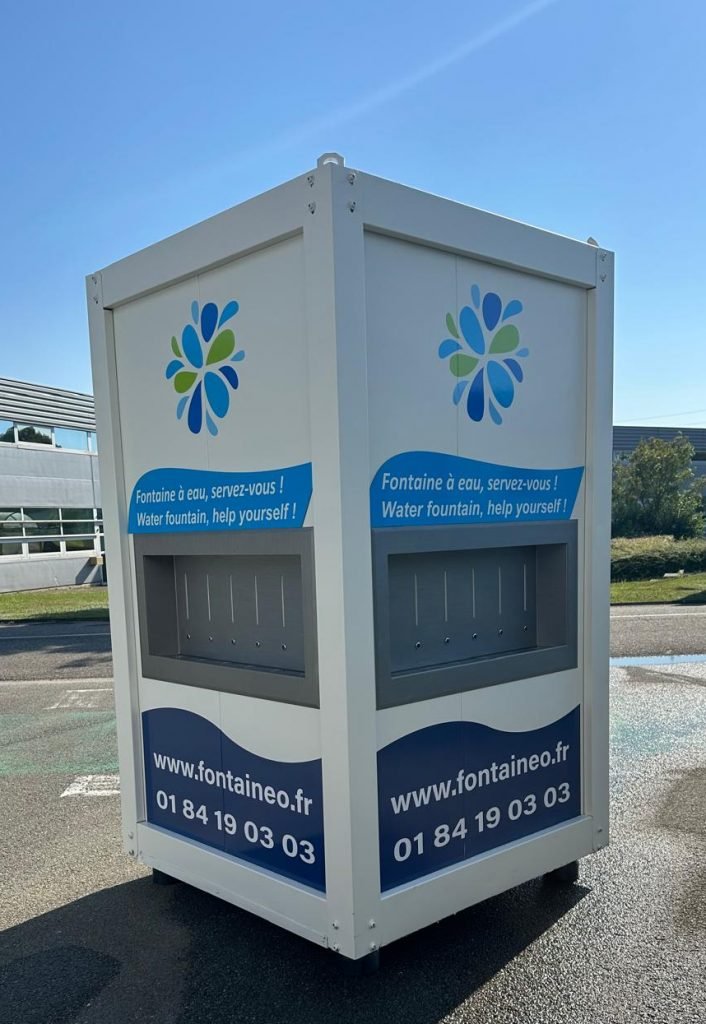 Fontaine à eau autonome sur un parking avec un logo bleu et vert, un lien vers un site Internet (www.fontaineo.eu) et un numéro de téléphone. Des panneaux en français et en anglais indiquent : « Fontaine à eau, servez-vous !.