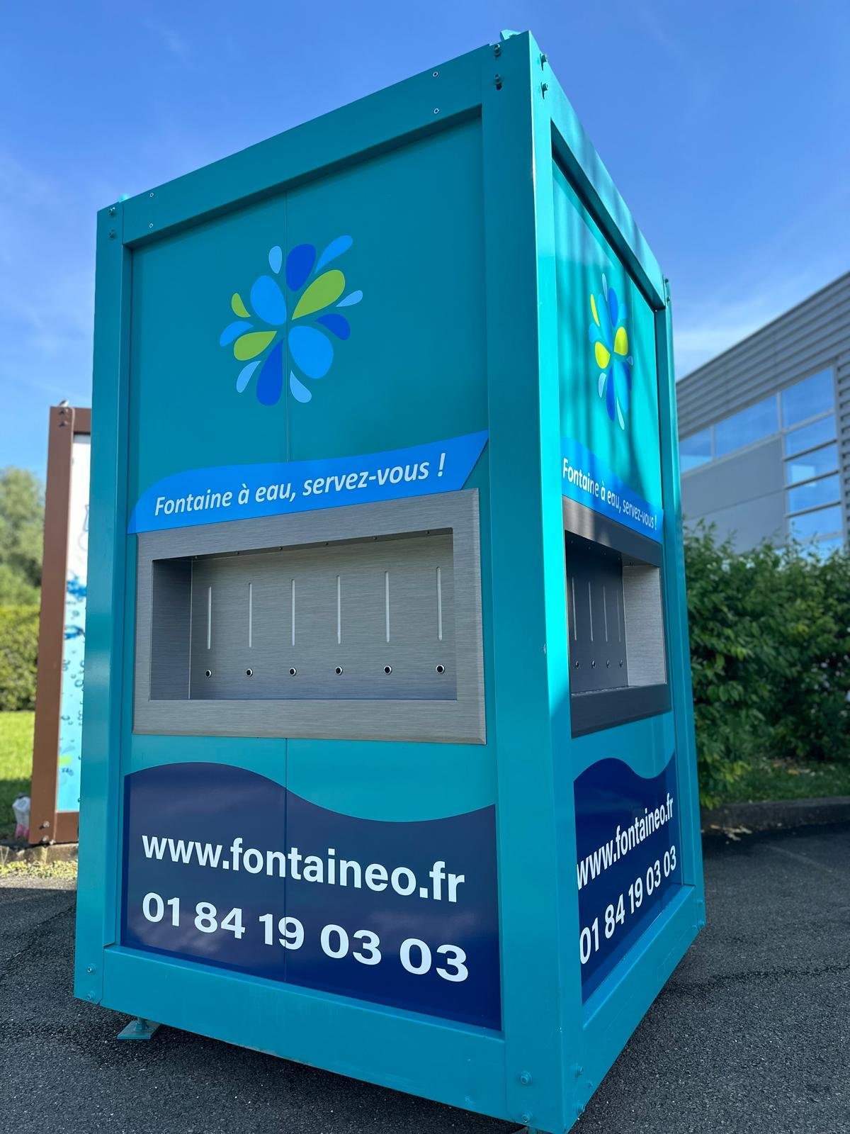 Un distributeur d'eau bleu sarcelle est présenté à l'extérieur, avec des logos représentant des gouttes d'eau bleues et vertes. Il est écrit « Fontaine à eau, servez-vous ! » et affiche un site Web et un numéro de téléphone. Un bâtiment et de la verdure sont visibles en arrière-plan.