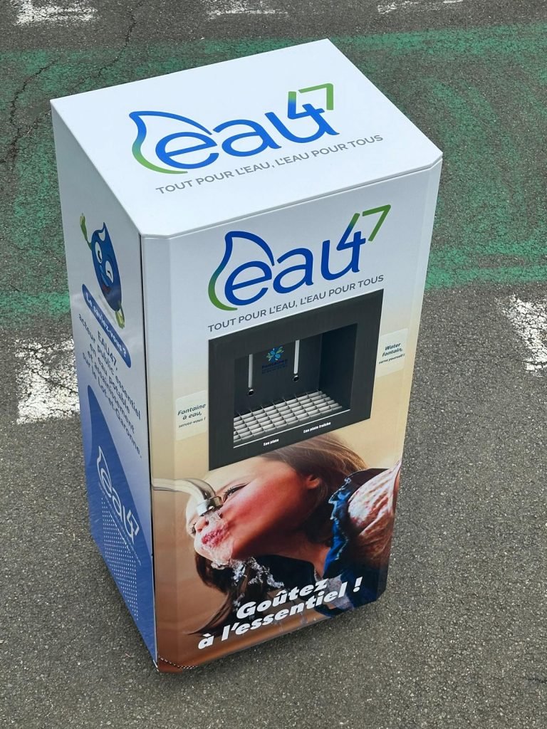 Un distributeur d'eau rectangulaire sur le trottoir, portant l'inscription « eau4 » et le slogan « Tout pour l'eau, l'eau pour tous ». Il présente l'image d'une personne buvant de l'eau et le texte « Goûtez à l'essentiel ! » sur le panneau avant.