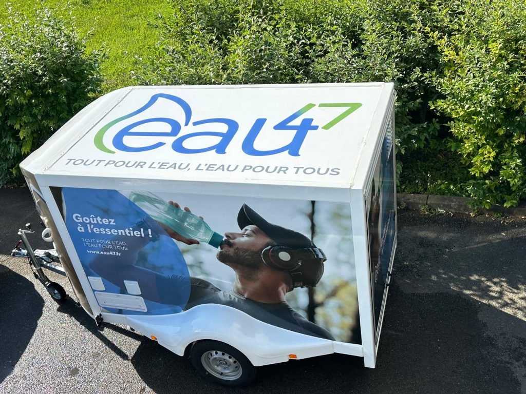 Une remorque blanche avec une grande publicité représentant un homme buvant de l'eau dans une bouteille. Les mots « eau 47 » et « Tout pour l'eau, l'eau pour tous » y sont inscrits. La remorque est garée sur une route avec de la verdure en arrière-plan.
