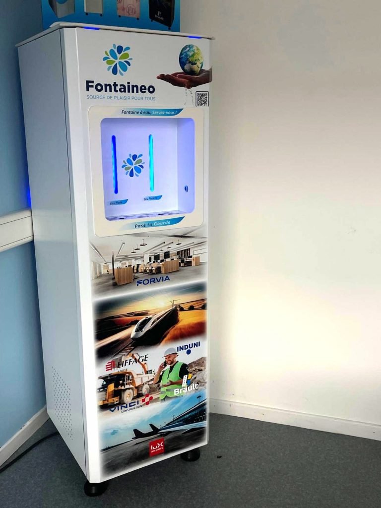 Un distributeur automatique blanc portant l'inscription « Fontaineo » est placé contre un mur. La machine est dotée d'un écran numérique en haut et de diverses publicités en bas. Elle repose sur quatre petites roues sur un sol gris.