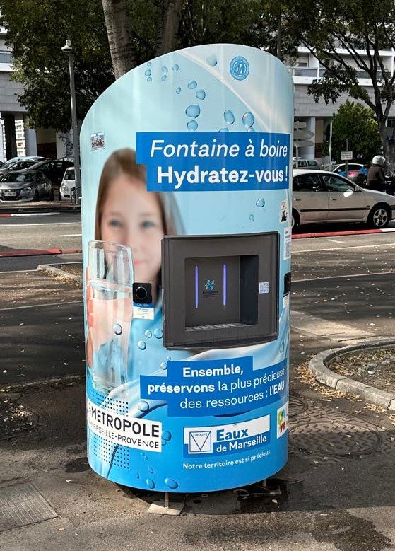 Fontaine à eau cylindrique avec un revêtement bleu affichant l'image d'une personne en train de boire de l'eau. Le texte en français encourage l'hydratation et la conservation de l'eau. Les logos de la Métropole Aix-Marseille-Provence et des Eaux de Marseille sont visibles.