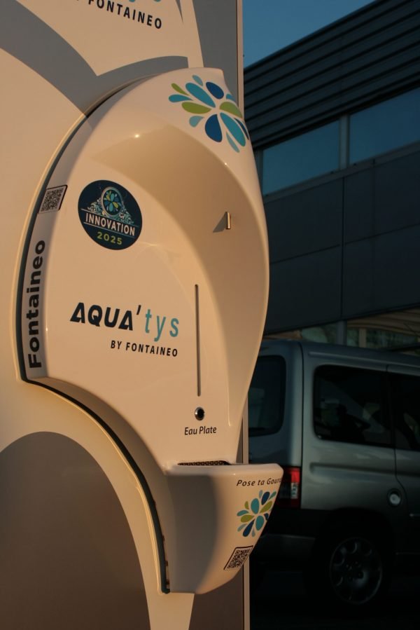 Un appareil blanc estampillé « AQUAtys by Fontaeneo » est fixé au mur. Il présente un design élégant et incurvé avec des motifs floraux bleus et verts. Sous l'étiquette, on peut lire « Eau Plate ». Un véhicule est partiellement visible en arrière-plan.