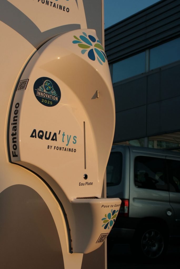 Un appareil blanc estampillé « AQUAtys by Fontaeneo » est fixé au mur. Il présente un design élégant et incurvé avec des motifs floraux bleus et verts. Sous l'étiquette, on peut lire « Eau Plate ». Un véhicule est partiellement visible en arrière-plan.