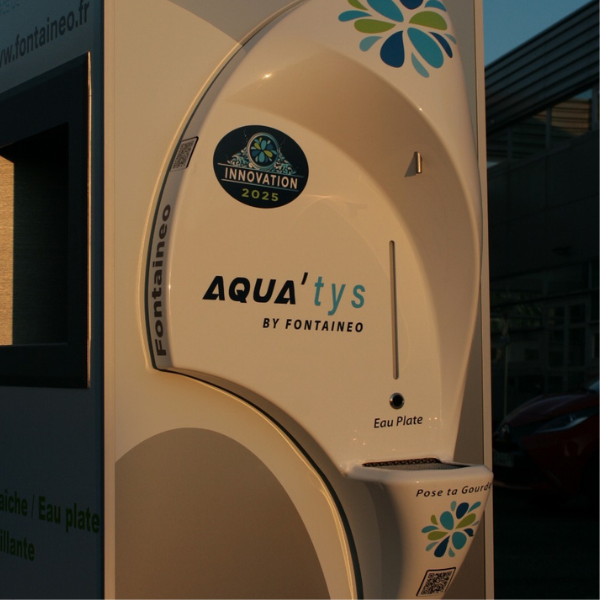 Close-up of an AQUA'tys water dispenser from Fontaigueo, with a rounded design and the inscription "Innovation 2025". It features slots labeled "Eau Plate" for dispensing still water, as well as the logo and website address.