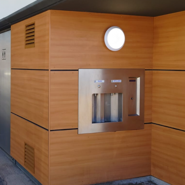 Station de remplissage murale pour bouteilles d'eau, équipée de deux distributeurs et de boutons étiquetés pour l'eau du robinet et l'eau gazeuse. L'appareil est installé sur un panneau extérieur en bois, surmonté d'un éclairage circulaire.