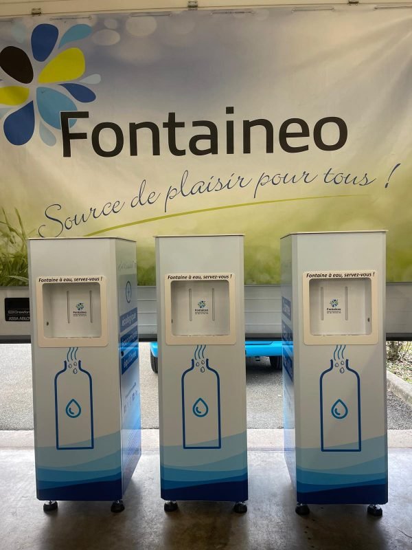 Trois fontaines à eau blanches et bleues sont alignées devant une bannière avec le logo et le nom « Fontaineo ». La bannière comprend le slogan « Source de plaisir pour tous ! » Les fontaines sont ornées d'une illustration de goutte d'eau et de bouteille bleue.