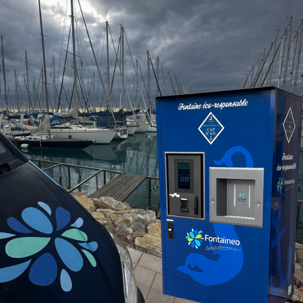 Un distributeur d'eau bleu, labélisé « Fontaineo éco-responsable », est situé près d'un port où naviguent des voiliers. Il se trouve à côté d'un véhicule sombre arborant un logo en forme de fleur. Le ciel est nuageux et un chemin de pierre longe l'eau.