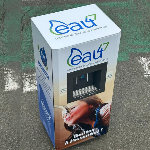 Un distributeur d'eau rectangulaire, baptisé « Eau4 », est installé sur un trottoir. Il arbore un logo et la photo d'une personne buvant de l'eau. Un texte en français est visible, encourageant l'accès à l'eau. Le distributeur dispose d'une zone de distribution à l'avant.