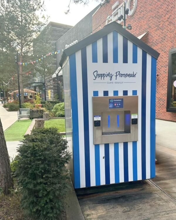 Un kiosque rayé bleu et blanc portant l'inscription "Shopping Promenade" se trouve sur un trottoir à côté d'arbres et d'arbustes, avec un distributeur d'eau potable intégré et des bancs visibles à l'arrière-plan.