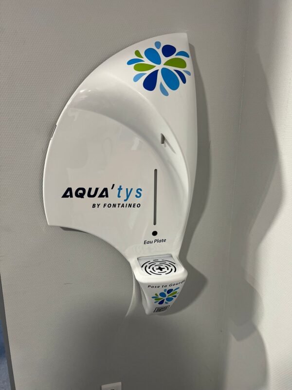 Un distributeur d'eau blanc moderne, étiqueté "AQUA'tys by FONTAINEO" avec un logo de gouttelettes bleues et vertes, est monté sur un mur gris au-dessus d'un porte-gobelet intégré. Le distributeur indique "Eau Plate" (eau plate).