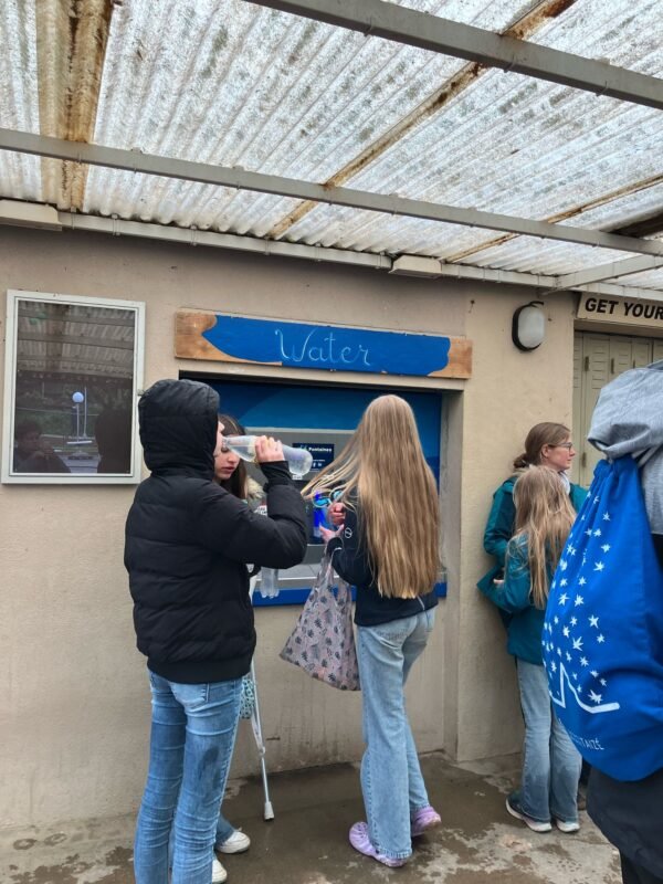 Plusieurs personnes, principalement des filles, se tiennent sous un abri près d'une fontaine murale étiquetée « Eau ». Une fille boit à la bouteille tandis que d'autres attendent ou discutent. Le temps semble pluvieux ou humide.