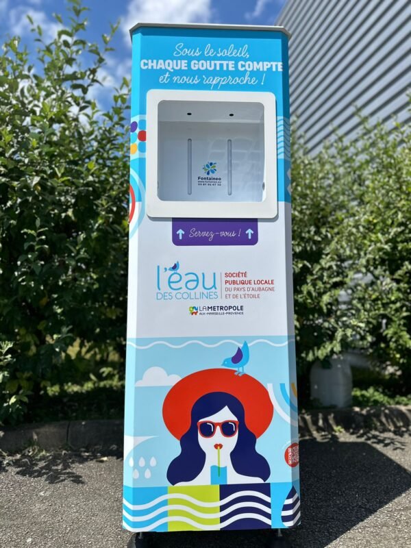 Distributeur d'eau public coloré représentant une femme portant des lunettes de soleil et un chapeau et buvant à la paille, avec l'inscription "l'eau des collines" et des instructions en français. Le distributeur est situé à l'extérieur, près d'un bâtiment et de verdure.