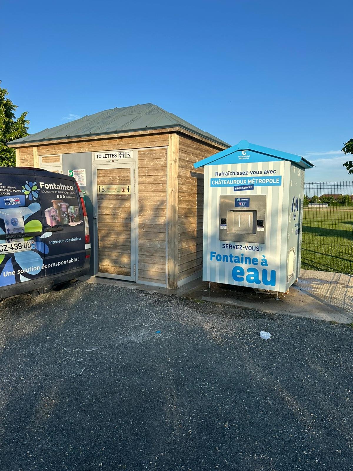 Un bâtiment en bois avec toilettes publiques et un distributeur d'eau bleu portant l'inscription « Fontaine à eau » sont situés côte à côte sur un espace extérieur pavé, sous un ciel dégagé. Une camionnette de marque est partiellement visible sur la gauche.