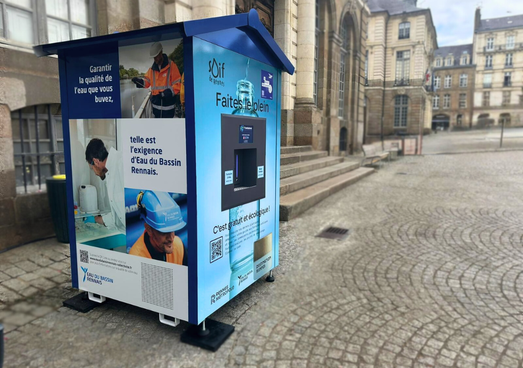 En una plaza adoquinada, cerca de edificios históricos, hay un dispensador público de agua de color azul. El quiosco muestra imágenes de personas bebiendo agua y llevando equipo de seguridad, así como un texto en francés sobre la calidad del agua.