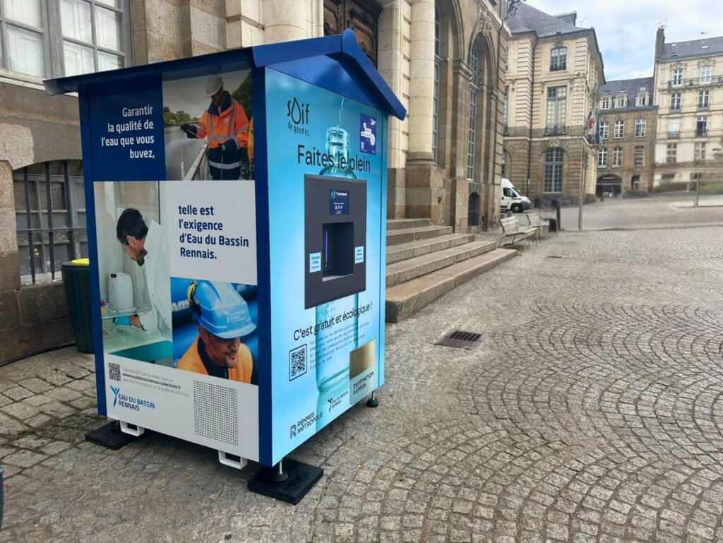 Un kiosque bleu et blanc de distribution d'eau publique se trouve dans une rue pavée de pierres, à proximité de bâtiments historiques. Le kiosque présente des graphiques et des photos d'information sur la qualité et la sécurité de l'eau.