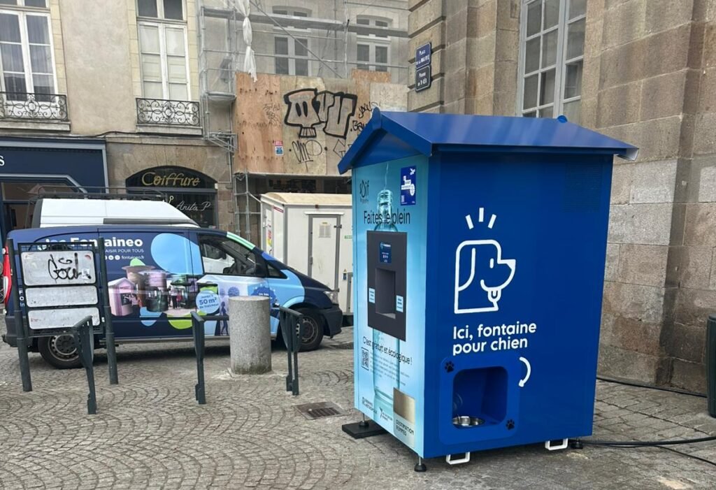 Une fontaine à eau pour chiens de couleur bleue se trouve dans une rue pavée, près d'une camionnette bleue. De vieux bâtiments et un échafaudage de construction sont visibles à l'arrière-plan. La fontaine est ornée d'une icône représentant un chien et d'un texte en français.