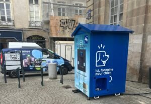 Une fontaine à eau pour chiens de couleur bleue se trouve dans une rue pavée, près d'une camionnette bleue. De vieux bâtiments et un échafaudage de construction sont visibles à l'arrière-plan. La fontaine est ornée d'une icône représentant un chien et d'un texte en français.