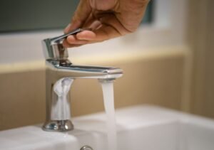 Une main ouvre un robinet argenté et de l'eau coule dans un lavabo blanc dans une salle de bains.
