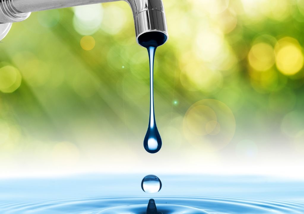 Gros plan d'un robinet en métal avec une goutte d'eau tombant dans une piscine en dessous, créant des ondulations, sur un fond flou vert et jaune.