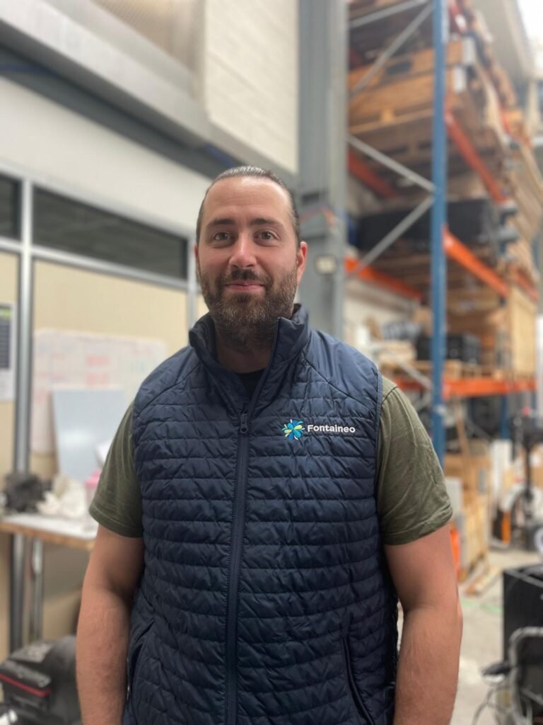 Un homme barbu, portant un gilet matelassé bleu foncé avec le logo "Fontaineo" sur un T-shirt vert, se tient à l'intérieur d'un entrepôt avec des étagères et divers équipements en arrière-plan.