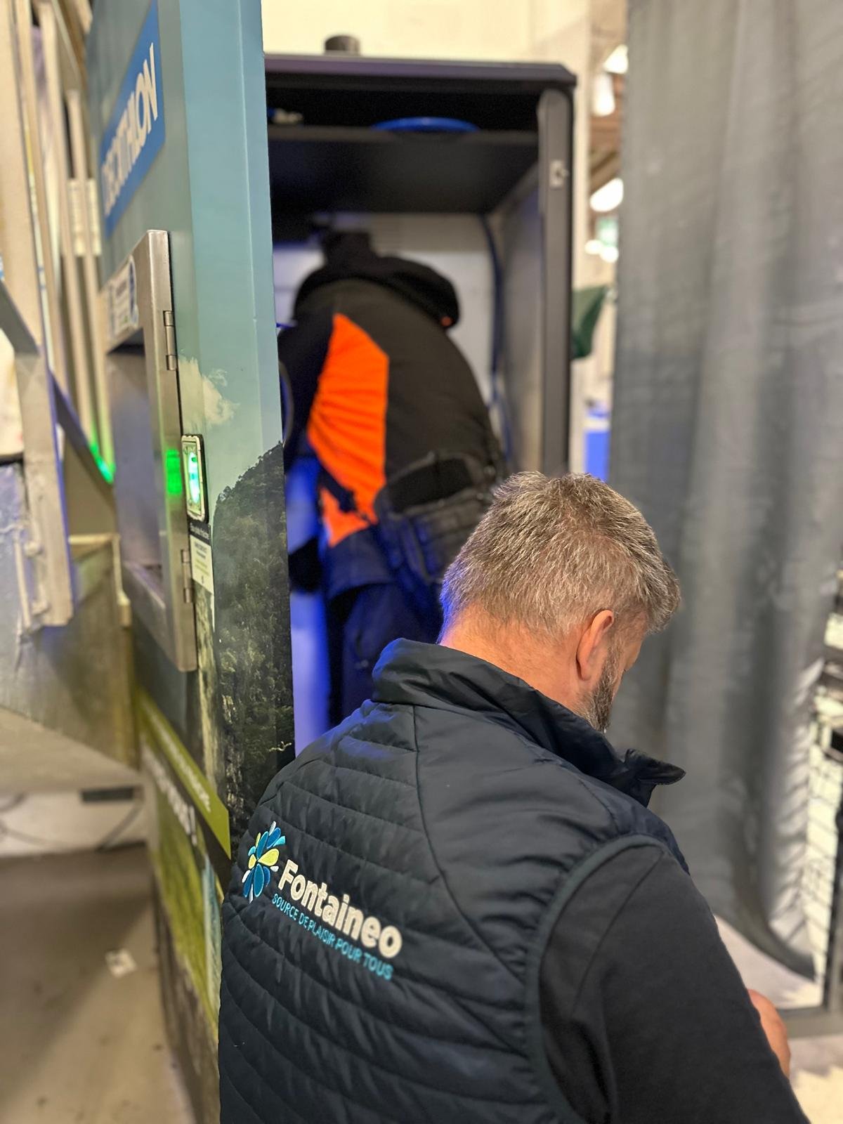 Une personne portant un gilet "Fontaineo" est agenouillée à côté d'une machine à distribuer de l'eau, tandis qu'une autre personne portant une veste orange et noire travaille à l'intérieur de la machine. La zone semble industrielle et divers équipements sont visibles.