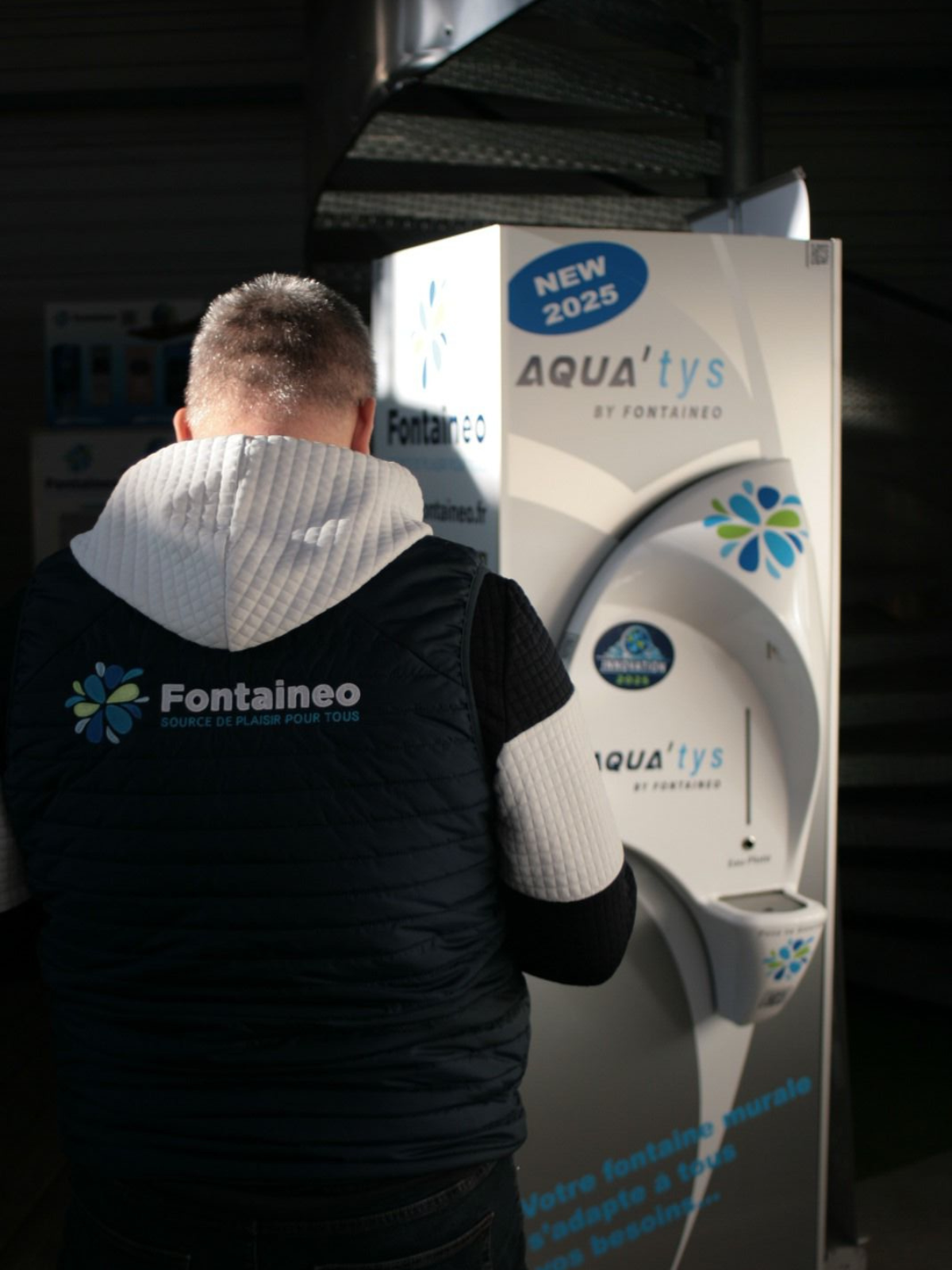 Une personne portant un gilet "Fontaineo" se tient devant un distributeur d'eau Aqua'tys portant l'étiquette "NEW 2025", partiellement éclairé par la lumière du soleil.