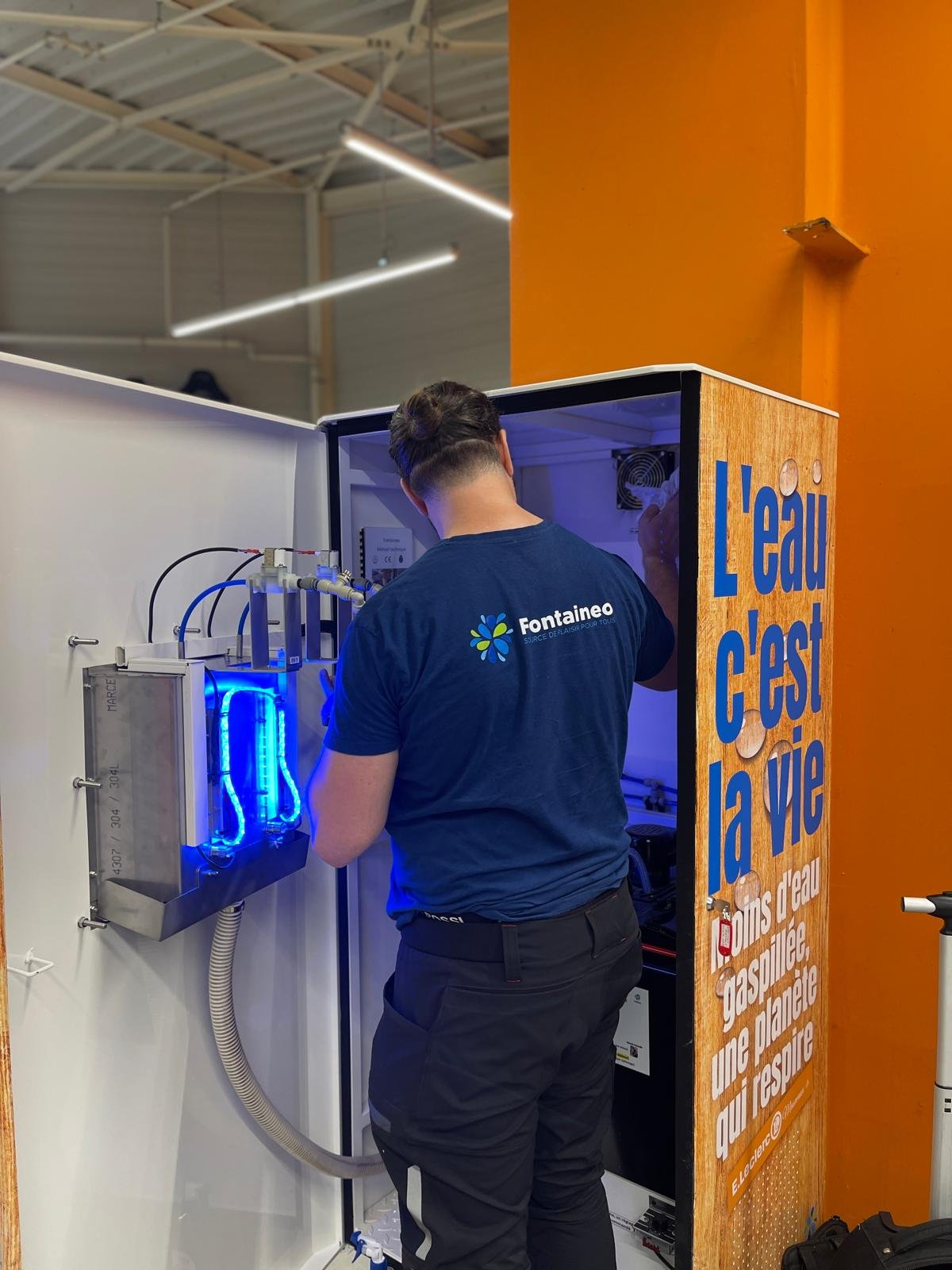 Un technicien portant une chemise Fontainéo travaille sur un équipement dont les composants sont éclairés en bleu à l'intérieur d'une machine ouverte. La machine porte le texte français "L'eau c'est la vie" et est adossée à un mur orange.
