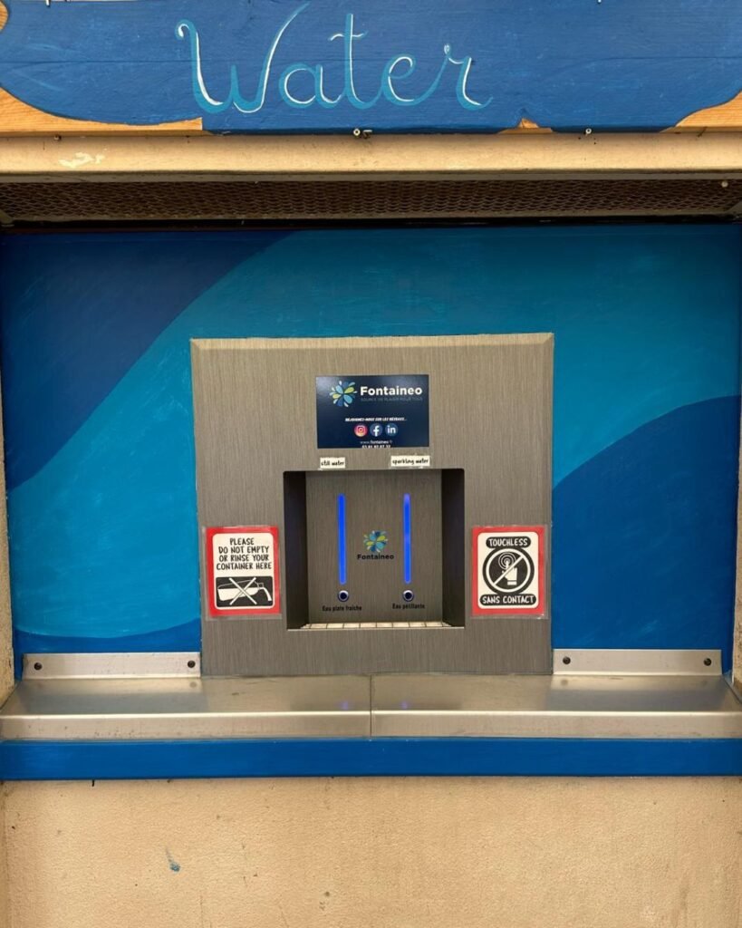 Station publique de remplissage d'eau avec deux becs, un logo "Fontaineo" et des panneaux indiquant "Uniquement pour le remplissage de récipients d'eau" et "Interdit de fumer, interdit aux animaux domestiques". Le fond est bleu avec le mot "Water" en haut.
