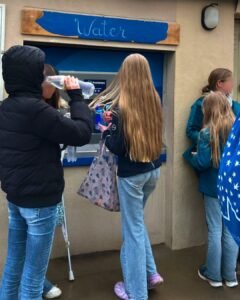Quatre jeunes se tiennent devant une station de remplissage d'eau ; l'un boit dans une bouteille, l'autre tient un sac réutilisable et l'un d'eux a des béquilles. Tous sont habillés de manière décontractée, avec des vestes et des jeans.