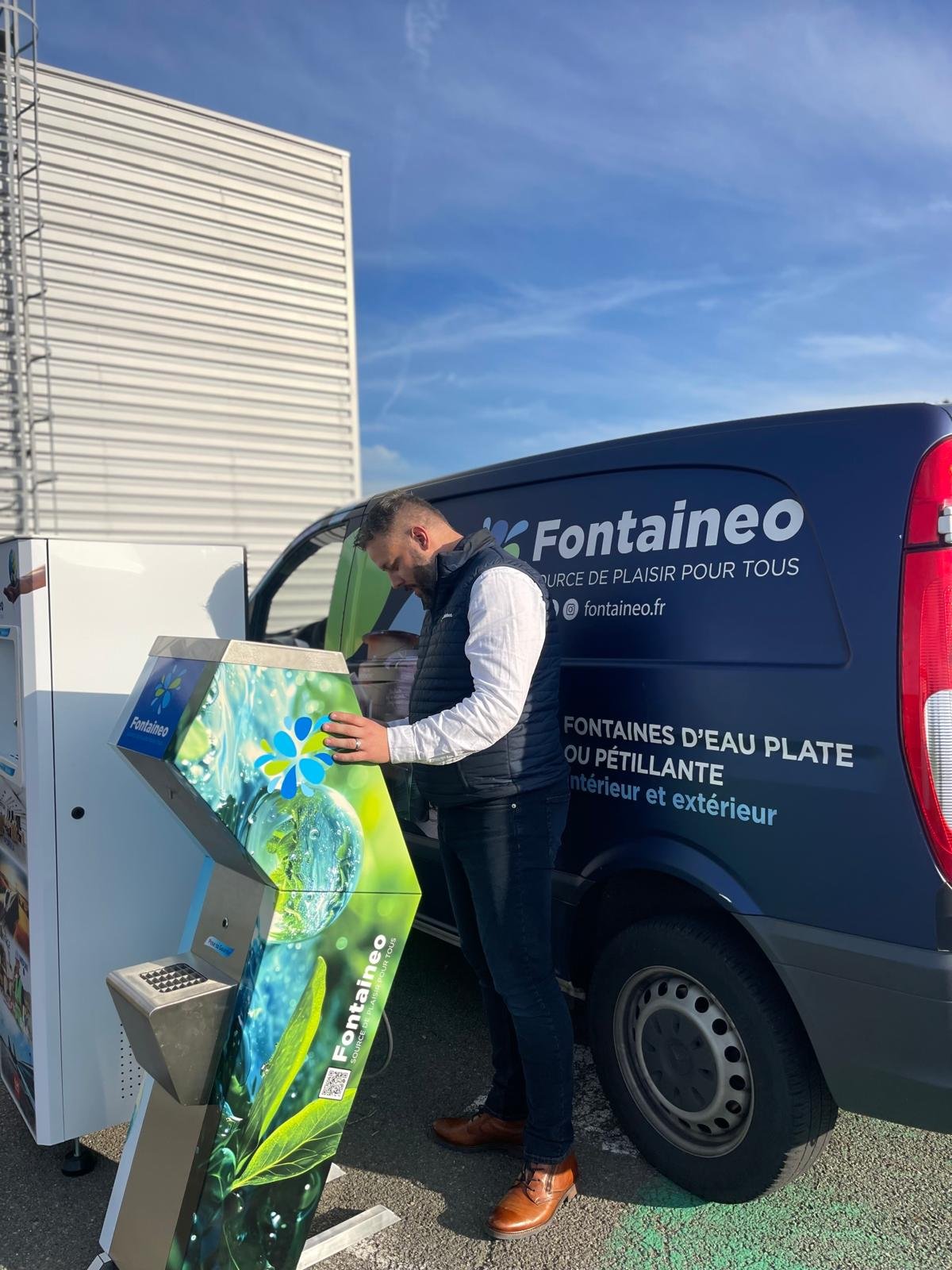 Un homme vêtu d'un gilet et d'un jean se tient à l'extérieur et utilise un distributeur d'eau Fontaineo à côté d'une camionnette bleue Fontaineo. La camionnette et le distributeur sont marqués du logo Fontaineo et d'images d'eau et de fleurs.