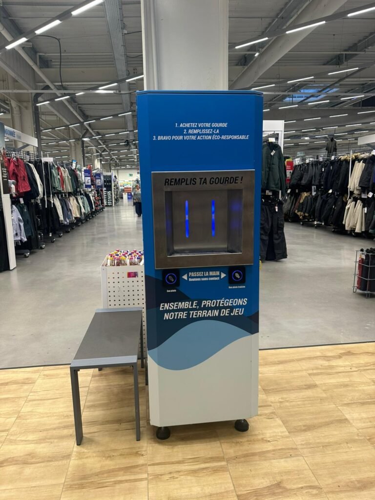 Station de remplissage d'eau dans un magasin de détail avec un panneau en français encourageant l'utilisation de bouteilles réutilisables. On aperçoit à l'arrière-plan des porte-vêtements et une petite table avec des articles.