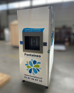 Un grand distributeur d'eau blanche étiqueté "Fontaineo" avec un écran numérique et deux boutons, situé dans un entrepôt. La machine affiche un logo floral bleu et vert et des informations de contact.