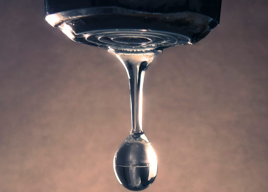 Vue rapprochée d'une goutte de liquide transparent qui pend et s'étire sur le bord d'une surface, sur un fond uni et légèrement éclairé.