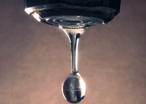 Vue rapprochée d'une goutte de liquide transparent qui pend et s'étire sur le bord d'une surface, sur un fond uni et légèrement éclairé.