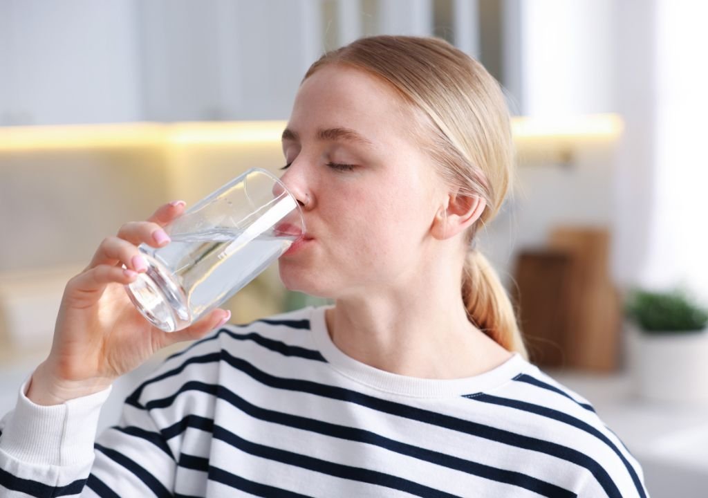 Une personne à la peau claire et aux cheveux blonds, vêtue d'une chemise à rayures noires et blanches, boit un verre d'eau dans un intérieur lumineux, en pensant aux quantités d'eau à boire par jour.