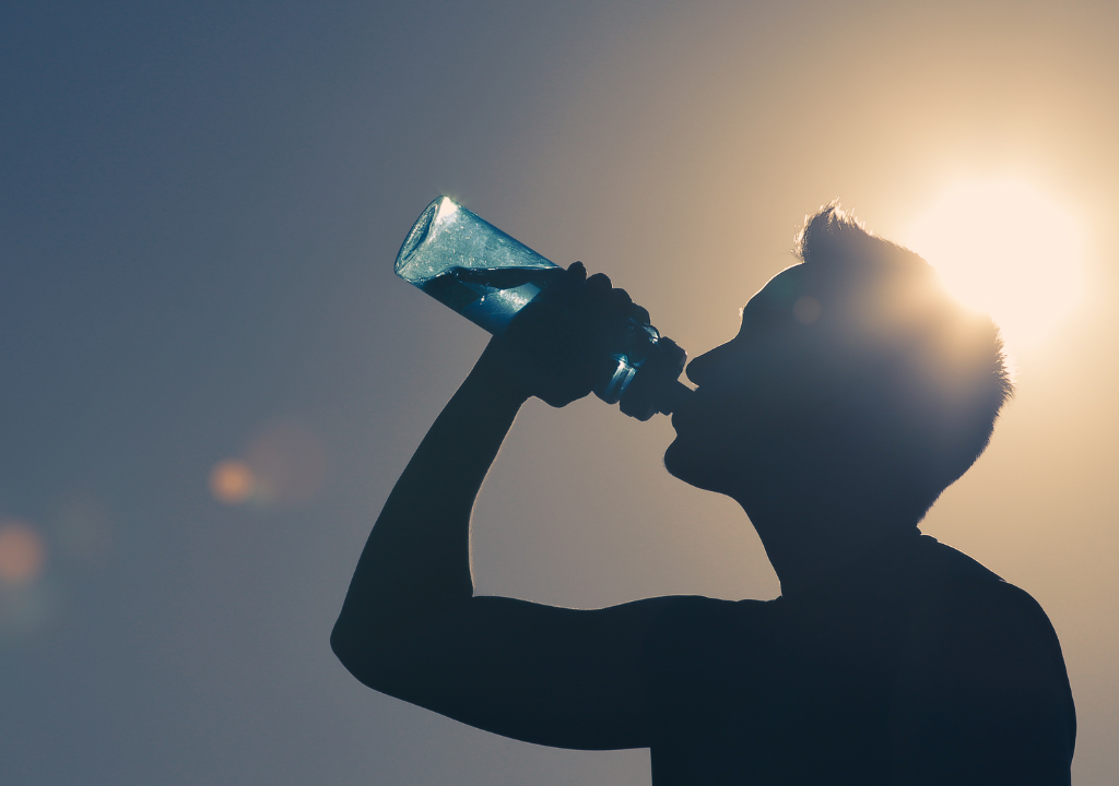 Une personne se découpe sur le soleil en buvant de l'eau potable dans une bouteille d'eau, avec un ciel clair en arrière-plan.