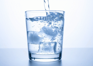 Un verre transparent rempli de glaçons repose sur une surface blanche tandis qu'on y verse de l'eau, ce qui provoque des bulles et des éclaboussures à l'intérieur du verre.