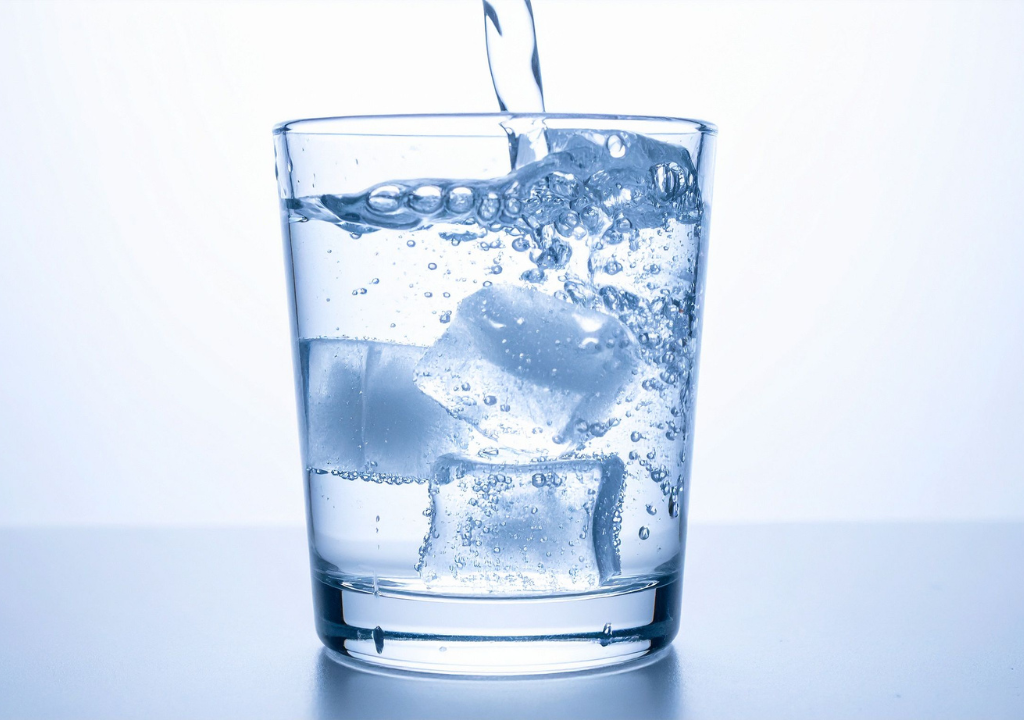 Un verre transparent rempli de glaçons repose sur une surface blanche tandis qu'on y verse de l'eau, ce qui provoque des bulles et des éclaboussures à l'intérieur du verre.