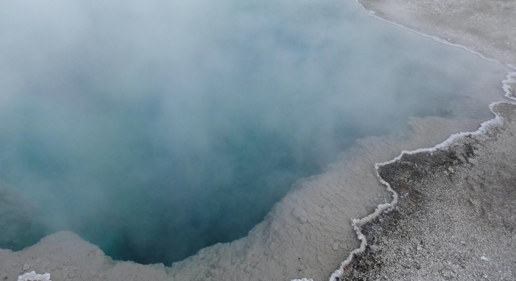 Une source d'eau chaude géothermique à l'eau bleue et vaporeuse jaillit d'un bassin profond, entouré de bords gris, rocheux et incrustés de minéraux. La vapeur masque une partie de l'eau et du terrain environnant.