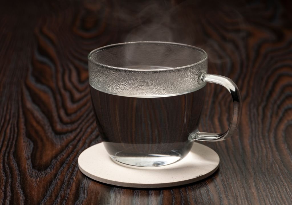 Une tasse en verre transparent remplie d'eau chaude est posée sur un sous-verre blanc surmontant une surface sombre en grain de bois. On peut voir de la vapeur s'échapper de l'eau.