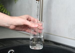 Une main tient un gobelet en verre sous un robinet, le remplissant d'eau. Le gobelet est marqué de lignes de mesure et de chiffres. La scène se déroule au-dessus d'un évier noir, avec un mur carrelé gris en arrière-plan.