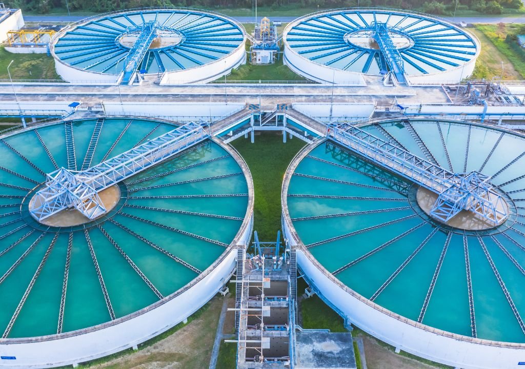 Vue aérienne d'une station d'épuration avec de grands bassins de décantation circulaires contenant de l'eau bleu-vert, des passerelles métalliques et des zones herbeuses - ce qui soulève la question suivante : peut-on boire une eau déminéralisée traitée dans de telles installations ?.