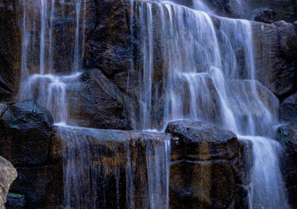 L'eau s'écoule sur plusieurs niveaux de roches sombres et humides dans une cascade. L'eau semble lisse et soyeuse, enrichie par les minéraux contenus dans l'eau, avec de la mousse verte sur les pierres. La scène est naturelle et tranquille.