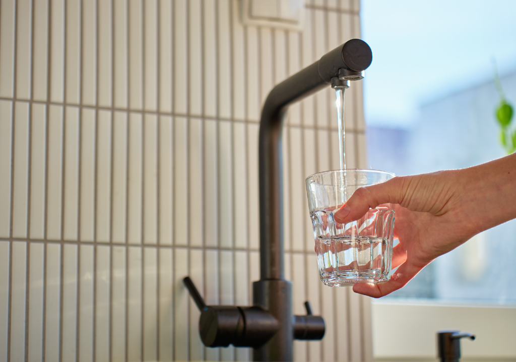 Une main tient un verre sous un robinet de cuisine qui coule, le remplissant d'eau. L'arrière-plan est composé de carreaux verticaux blancs et d'une partie d'une fenêtre où l'on voit la lumière du jour.