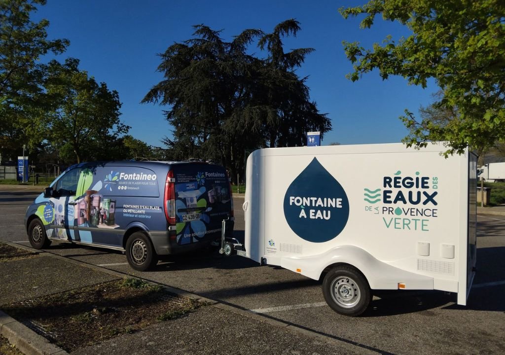 Une camionnette portant l'inscription "Fontaineo" est garée et tire une remorque blanche portant l'inscription "Fontaine à Eau" et "Régie des Eaux de Provence Verte" dans un cadre extérieur ensoleillé avec des arbres et un sol pavé.