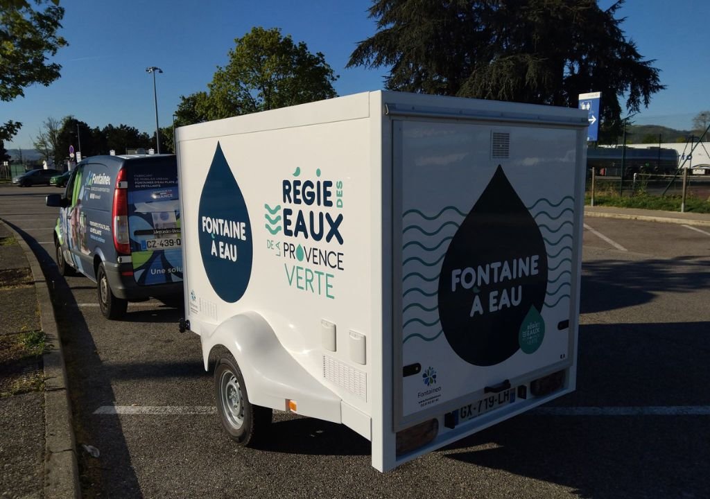 Une remorque blanche avec de grandes gouttes d'eau et les textes français "Fontaine à Eau" et "Régie des Eaux Provence Verte" est garée sur un terrain à côté d'une camionnette. Des arbres et un ciel bleu sont visibles en arrière-plan.