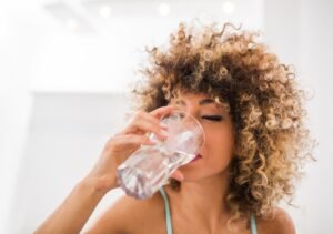 Une personne aux cheveux bouclés boit de l'eau dans un verre transparent, en intérieur, sur un fond lumineux et flou.